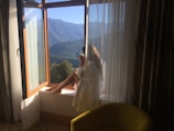 A person relaxes at an open window, gazing at a scenic mountain landscape. The room is softly lit, with sheer curtains allowing gentle sunlight to filter in. A yellow chair is positioned nearby, adding a touch of color to the cozy, peaceful space.