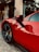 A close-up of a sleek, red sports car parked on cobblestone pavement in front of a building with a sign reading 'Hotel de Paris'. The car's door and wheel are prominently featured, with the iconic prancing horse emblem visible on the fender. A small tree is part of the background next to the hotel entrance.
