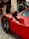 A close-up of a sleek, red sports car parked on cobblestone pavement in front of a building with a sign reading 'Hotel de Paris'. The car's door and wheel are prominently featured, with the iconic prancing horse emblem visible on the fender. A small tree is part of the background next to the hotel entrance.