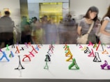 A gallery space filled with a collection of small, colorful abstract sculptures displayed on a white table. The sculptures are made of various colors like green, red, pink, purple, and orange. People are gathered in the background, some in motion blur, indicating movement and a busy setting.