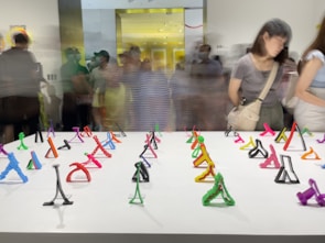 A gallery space filled with a collection of small, colorful abstract sculptures displayed on a white table. The sculptures are made of various colors like green, red, pink, purple, and orange. People are gathered in the background, some in motion blur, indicating movement and a busy setting.