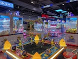 Brightly lit arcade filled with various game machines, including claw machines with stuffed toys, a ticket redemption game, and colorful decorative lighting. The environment is lively and playful, with numerous arcade game consoles lining the floor.
