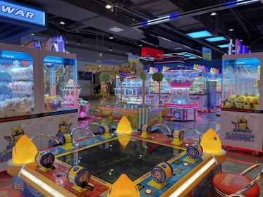 Brightly lit arcade filled with various game machines, including claw machines with stuffed toys, a ticket redemption game, and colorful decorative lighting. The environment is lively and playful, with numerous arcade game consoles lining the floor.