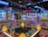 Brightly lit arcade filled with various game machines, including claw machines with stuffed toys, a ticket redemption game, and colorful decorative lighting. The environment is lively and playful, with numerous arcade game consoles lining the floor.