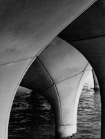 An architectural image showing the underside of a bridge with large, curving concrete arches. The surface of a body of water is visible in the background, adding texture to the scene.