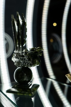 A detailed sculpture of a ship made of dark green material, displayed on a reflective surface with bright, circular LED lights in the background.