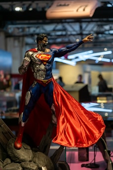 A detailed model of a superhero, featuring parts with exposed mechanical components and a red and blue costume. The figure is standing in a confident pose with an extended right arm and a flowing red cape. The background is blurred, indicating an indoor setting with people and structures that resemble a convention or exhibit environment.