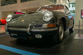 A classic Porsche car displayed in a museum setting. The vehicle features a sleek, dark body with shiny chrome elements and round headlights. The background includes a modern interior with subtle lighting and another red car partially visible. The floor has a glossy finish, enhancing the car's polished appearance.