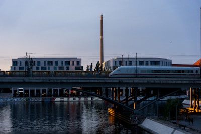 A sleek white passenger train travels on an elevated railway track over a calm waterway, with modern white buildings in the background. The reflection of the train and the bridge appears in the water. The sky is a soothing shade of blue as dusk approaches, and there are street lights and some greenery visible.