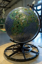 A large globe constructed from various electronic components, including circuit boards and microchips, displayed on a stand. The globe is intricately designed to resemble a map, with green and blue hues representing land and sea, respectively. The setting is an industrial or exhibition space with visible metallic beams and a large window in the background.