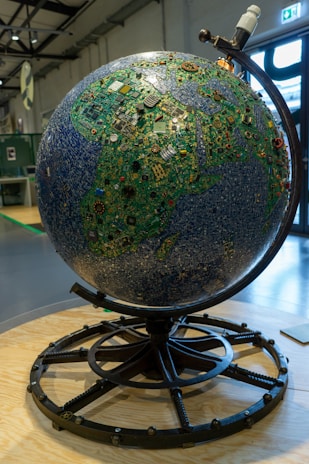 A large globe constructed from various electronic components, including circuit boards and microchips, displayed on a stand. The globe is intricately designed to resemble a map, with green and blue hues representing land and sea, respectively. The setting is an industrial or exhibition space with visible metallic beams and a large window in the background.