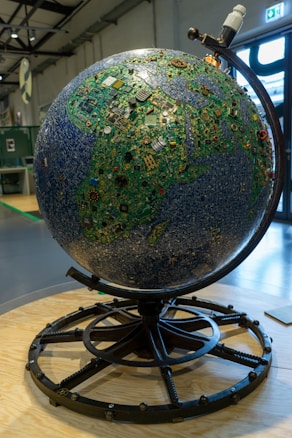 A large globe constructed from various electronic components, including circuit boards and microchips, displayed on a stand. The globe is intricately designed to resemble a map, with green and blue hues representing land and sea, respectively. The setting is an industrial or exhibition space with visible metallic beams and a large window in the background.