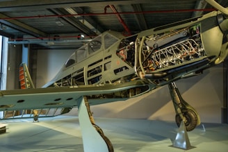 A partially restored vintage aircraft on display, with exposed internal components such as the engine and fuselage structure, showcased in a museum-like setting. The exterior paint is faded, indicating its age and historical significance. The surrounding environment includes industrial features like metal beams and subdued lighting.