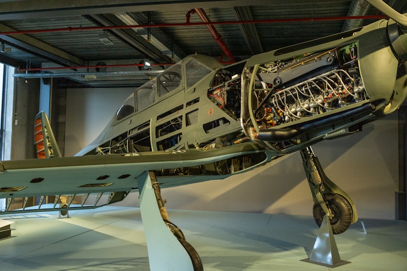 A partially restored vintage aircraft on display, with exposed internal components such as the engine and fuselage structure, showcased in a museum-like setting. The exterior paint is faded, indicating its age and historical significance. The surrounding environment includes industrial features like metal beams and subdued lighting.
