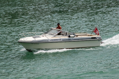 A motorboat with a red flag depicting a white cross navigates through clear green waters. The boat is cream-colored with a person seated inside wearing a hat and a red shirt, steering the vessel. The water around the boat is rippling due to its movement, and the scene is brightly lit, suggesting daylight.
