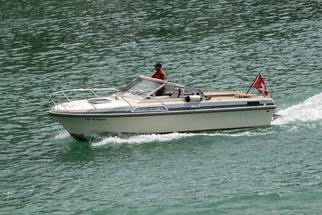 A motorboat with a red flag depicting a white cross navigates through clear green waters. The boat is cream-colored with a person seated inside wearing a hat and a red shirt, steering the vessel. The water around the boat is rippling due to its movement, and the scene is brightly lit, suggesting daylight.