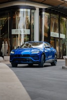 A sleek blue luxury SUV is parked in front of a high-end fashion store with large glass windows. The store has mannequins in the display and a sign with a stylish logo. The setting appears to be urban and upscale.