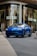 A sleek blue luxury SUV is parked in front of a high-end fashion store with large glass windows. The store has mannequins in the display and a sign with a stylish logo. The setting appears to be urban and upscale.