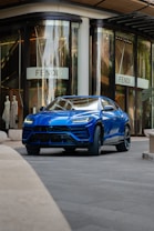 A sleek blue luxury SUV is parked in front of a high-end fashion store with large glass windows. The store has mannequins in the display and a sign with a stylish logo. The setting appears to be urban and upscale.