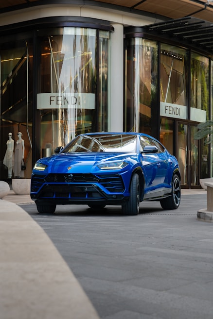 A sleek blue luxury SUV is parked in front of a high-end fashion store with large glass windows. The store has mannequins in the display and a sign with a stylish logo. The setting appears to be urban and upscale.