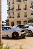 A luxurious sports car with a sleek design is parked in front of an elegant classical building with ornate balconies. The car features black rims and a prominent rear spoiler. The streetlight and trimmed bushes enhance the opulent setting.