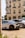 A luxurious sports car with a sleek design is parked in front of an elegant classical building with ornate balconies. The car features black rims and a prominent rear spoiler. The streetlight and trimmed bushes enhance the opulent setting.
