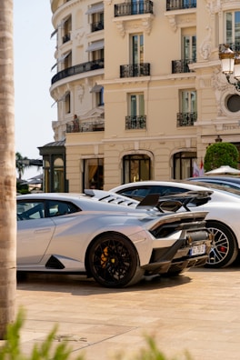 A luxurious sports car with a sleek design is parked in front of an elegant classical building with ornate balconies. The car features black rims and a prominent rear spoiler. The streetlight and trimmed bushes enhance the opulent setting.