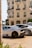 A luxurious sports car with a sleek design is parked in front of an elegant classical building with ornate balconies. The car features black rims and a prominent rear spoiler. The streetlight and trimmed bushes enhance the opulent setting.