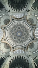 An intricate and ornate dome ceiling, featuring elaborate geometric and floral patterns with calligraphy. The design incorporates a blend of colors and textures, with detailed craftsmanship and symmetry. The architectural details showcase elements typical of a mosque, with repeating arches and intricate motifs.
