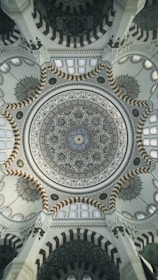 An intricate and ornate dome ceiling, featuring elaborate geometric and floral patterns with calligraphy. The design incorporates a blend of colors and textures, with detailed craftsmanship and symmetry. The architectural details showcase elements typical of a mosque, with repeating arches and intricate motifs.