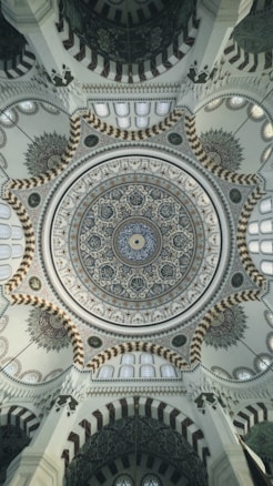 An intricate and ornate dome ceiling, featuring elaborate geometric and floral patterns with calligraphy. The design incorporates a blend of colors and textures, with detailed craftsmanship and symmetry. The architectural details showcase elements typical of a mosque, with repeating arches and intricate motifs.