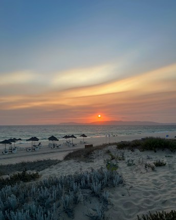 A serene beach with gentle waves lapping against the shore as the sun sets on the horizon. The sky is a blend of soft blues and warm oranges. There are sun loungers and umbrellas on the sand, with sparse greenery in the foreground.