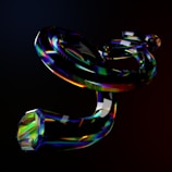 A dark, abstract visual featuring intertwined, glossy, and reflective shapes with rainbow-like colors. The objects resemble twisted ribbons or tubes, displaying a prismatic effect, against a dark background.