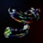 A dark, abstract visual featuring intertwined, glossy, and reflective shapes with rainbow-like colors. The objects resemble twisted ribbons or tubes, displaying a prismatic effect, against a dark background.