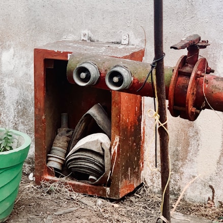 An old fire hose cabinet with a rusty exterior is attached to a red fire hydrant. The cabinet contains neatly coiled fire hoses. Surrounding the setup is a weathered concrete wall, and a green plant pot is visible nearby.