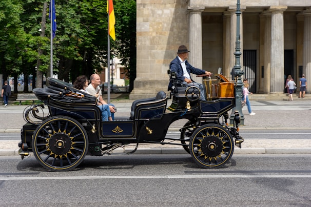 A vintage, open-top automobile is traveling along a city street. The vehicle is dark in color with gold accents and large spoked wheels. A driver wearing a hat operates the car, while two passengers sit in the back, enjoying the ride. In the background, there are people walking and a building with columns.