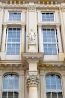 Detailed architectural facade featuring large windows with white frames and intricate decorative elements. A prominent central statue of a classical figure stands above a decorative corinthian column. The facade is ornate with various classical adornments including pilasters and cornices.