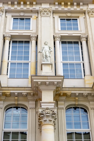 Detailed architectural facade featuring large windows with white frames and intricate decorative elements. A prominent central statue of a classical figure stands above a decorative corinthian column. The facade is ornate with various classical adornments including pilasters and cornices.