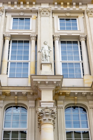 Detailed architectural facade featuring large windows with white frames and intricate decorative elements. A prominent central statue of a classical figure stands above a decorative corinthian column. The facade is ornate with various classical adornments including pilasters and cornices.