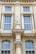 Detailed architectural facade featuring large windows with white frames and intricate decorative elements. A prominent central statue of a classical figure stands above a decorative corinthian column. The facade is ornate with various classical adornments including pilasters and cornices.