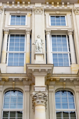 Detailed architectural facade featuring large windows with white frames and intricate decorative elements. A prominent central statue of a classical figure stands above a decorative corinthian column. The facade is ornate with various classical adornments including pilasters and cornices.