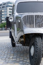 A vintage silver truck is parked on a cobblestone surface with modern buildings in the background. The truck has a prominent round headlight and a distinctive vertical grille at the front. The tires appear rugged and suitable for off-road conditions.