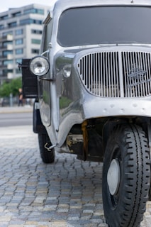 A vintage silver truck is parked on a cobblestone surface with modern buildings in the background. The truck has a prominent round headlight and a distinctive vertical grille at the front. The tires appear rugged and suitable for off-road conditions.