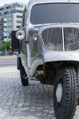 A vintage silver truck is parked on a cobblestone surface with modern buildings in the background. The truck has a prominent round headlight and a distinctive vertical grille at the front. The tires appear rugged and suitable for off-road conditions.