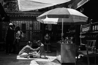 In a shaded area with an ornate backdrop, a person is receiving a massage or some form of care while sitting on a mat. Nearby stands a large umbrella providing extra shade. A group of people is gathered in the background, absorbed in conversation. The scene feels intimate and focused, with elements of daily life and cultural significance present.