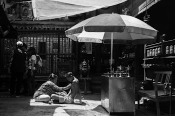 In a shaded area with an ornate backdrop, a person is receiving a massage or some form of care while sitting on a mat. Nearby stands a large umbrella providing extra shade. A group of people is gathered in the background, absorbed in conversation. The scene feels intimate and focused, with elements of daily life and cultural significance present.