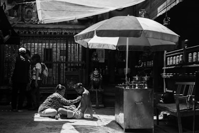 In a shaded area with an ornate backdrop, a person is receiving a massage or some form of care while sitting on a mat. Nearby stands a large umbrella providing extra shade. A group of people is gathered in the background, absorbed in conversation. The scene feels intimate and focused, with elements of daily life and cultural significance present.