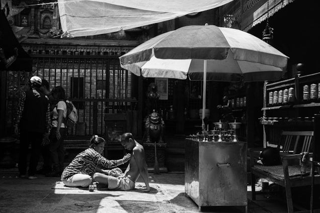 In a shaded area with an ornate backdrop, a person is receiving a massage or some form of care while sitting on a mat. Nearby stands a large umbrella providing extra shade. A group of people is gathered in the background, absorbed in conversation. The scene feels intimate and focused, with elements of daily life and cultural significance present.