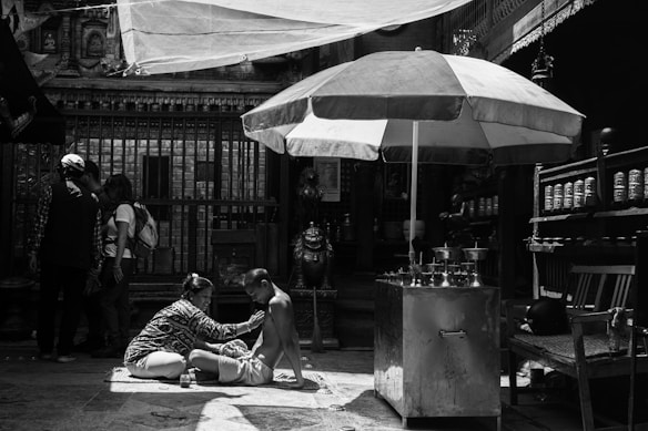 In a shaded area with an ornate backdrop, a person is receiving a massage or some form of care while sitting on a mat. Nearby stands a large umbrella providing extra shade. A group of people is gathered in the background, absorbed in conversation. The scene feels intimate and focused, with elements of daily life and cultural significance present.
