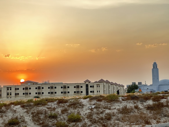 A sunset scene with the sun partially visible on the horizon, casting an orange glow across the sky. Below the sky, a modern building complex with a white facade and a tower with a distinctive design is situated. In the foreground, there is a sandy area with sparse vegetation.